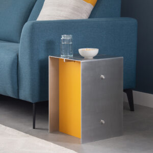 Photo of Core aluminium side table. The aluminium is folded around a warm yellow inside. On the table are a glass of water and a small bowl. The side table stands next to a blue couch with a light grey and yellow pillow.